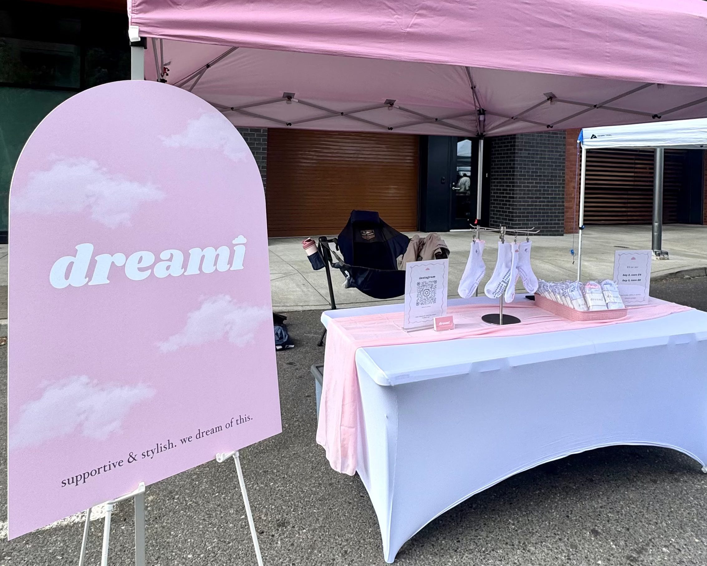 A pink canopy tent and matching pink sign stand on a gray paved outdoor area. A white-covered table with pink trim sits under the canopy, holding small display items.